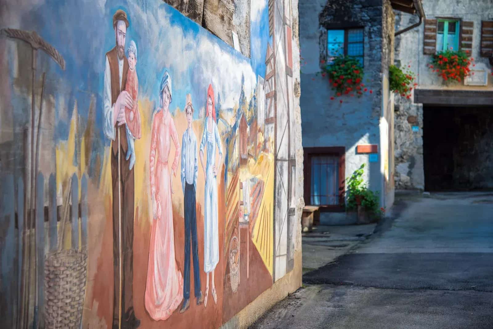 A painted mural of rural life on a stone house along a quiet alpine village lane