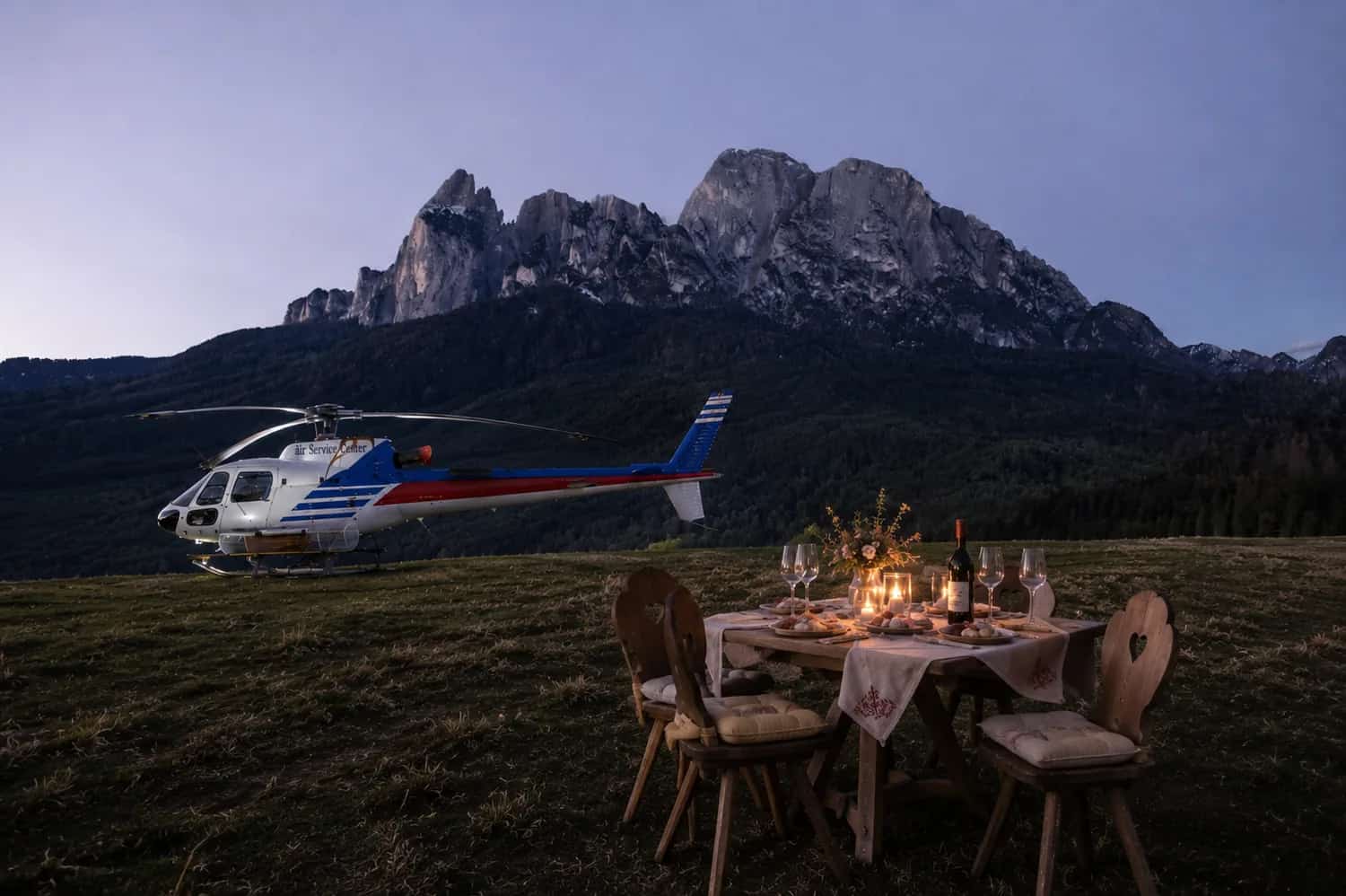 A private candlelit dinner laid in a meadow at dusk beside a helicopter, the Dolomites behind